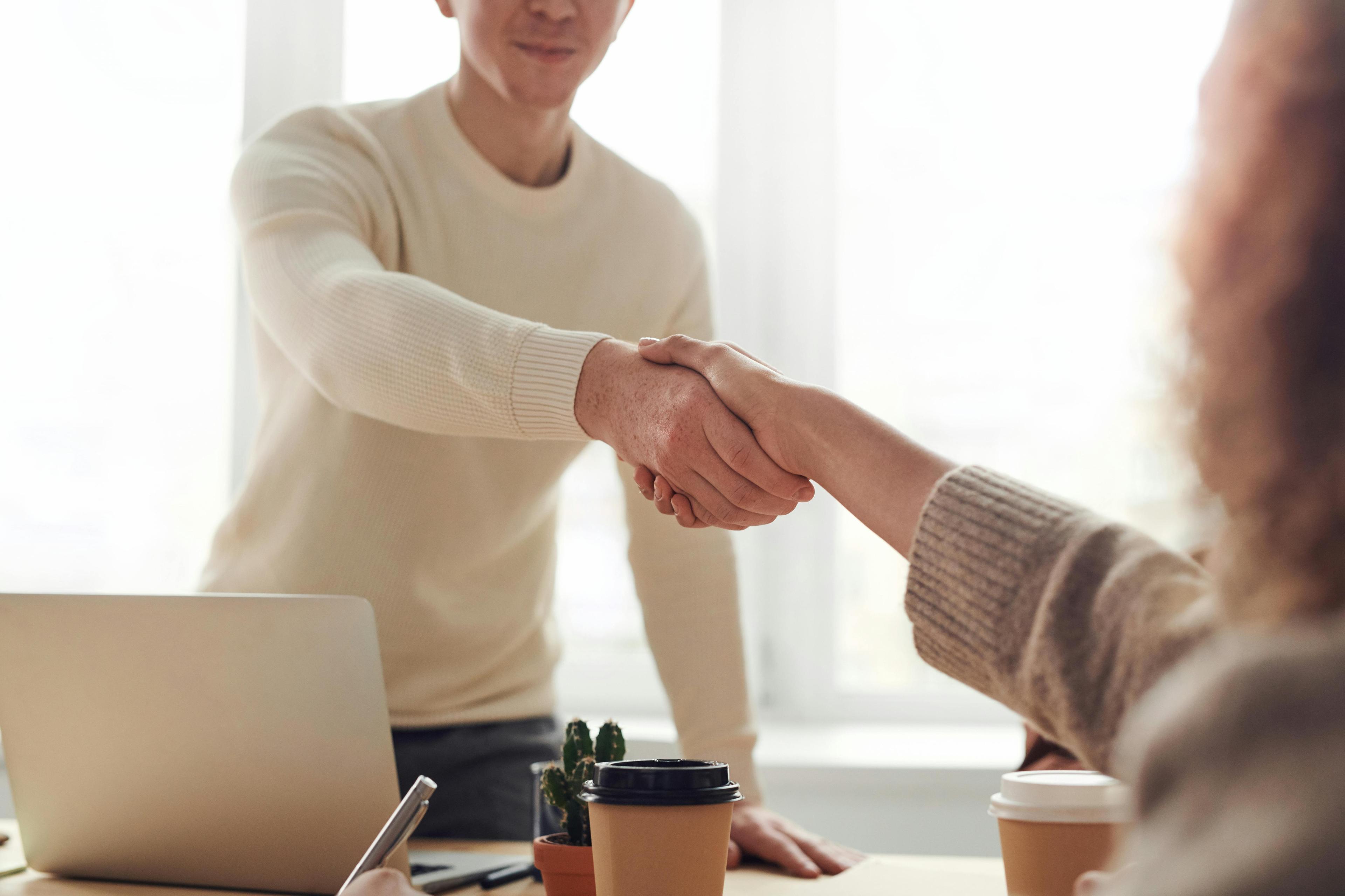 Man shakes hand of a woman. On the table are a laptop and 2 coffee to go.